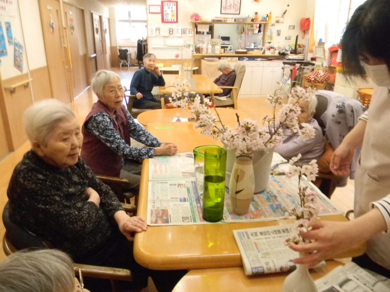 入居者と職員が共用スペースで新聞と花を囲んで交流している風景
